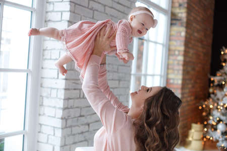 Cute little girl and her beautiful mother, dressed in pink dresses, spend time playing together indoors against the background of an elegant Christmas tree with golden garlands. Home portrait of mother and baby on the night before Christmas.の写真素材