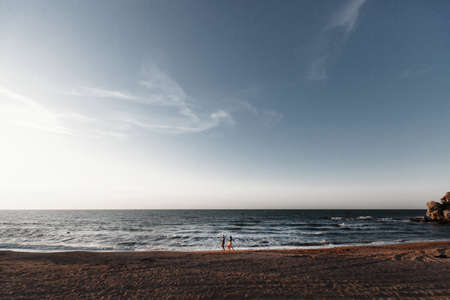 Couple in love at dawn by the sea. Honeymoon trip. Man and woman traveling. Happy couple by the sea view from the back. Man and woman holding hands. Couple in love on vacation. Follow me Couple on a rocky beach of blue ocean on a sunny dayの写真素材