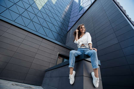 A brunette woman with long hair in fashionable in blue jeans and a white shirt posing outdoors. Fashion photo concept. Woman near business center. Portrait of a brunette woman. Beautiful female business center worker near her officeの写真素材