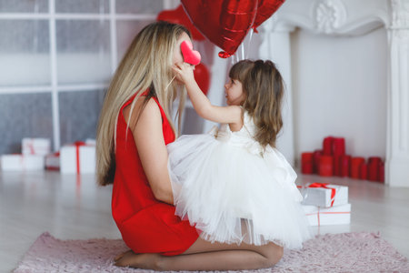 Mom and little daughter.Family portrait. Mother's day, love, family parenthood, childhood concept.Valentine's day - a young mother and little daughter in a room on the floor with gifts on the background of red balloons in the shape of a heartの写真素材
