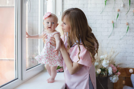 Mother and child on a white bed. Mom and baby girl in a sundress playing in sunny bedroom. Parent...