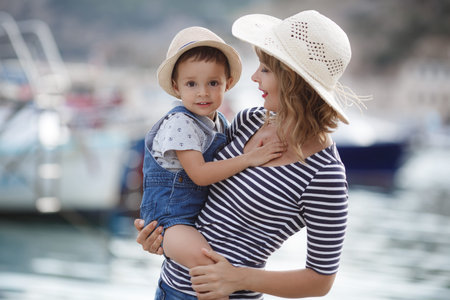 Mother and little son spend time outdoors in the summer together, sitting on a pier near boats and boats.Young mother plays with her baby on the promenade.Summer vacation for two by the sea. Mother and son at the resort together.の写真素材