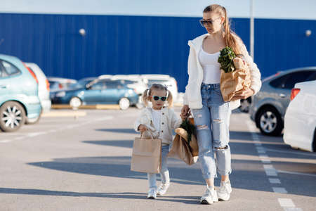 Mom and little daughter with ponytails, dressed in white T-shirts and blue jeans posing in the background of the parking lot near the supermarket with packages in hand. Mother and daughter shopping outdoors.の写真素材