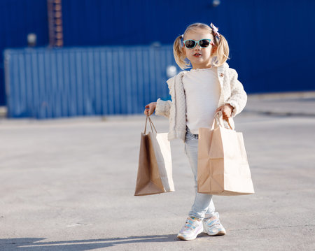 Cheerful baby with purchases. Little girl alone in spring outdoors with paper bags for shopping.Pretty girl, blonde in sun glasses posing alone on the street with shopping bags.の写真素材