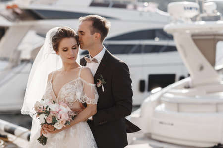 A pair of lovers walk on the pier near the yachts.Young couple in love outdoors in the summer, embracing on a pier on the background of beautiful white yachts and calm sea.A wonderful couple of newlyweds are walking along the pier by the seaの写真素材