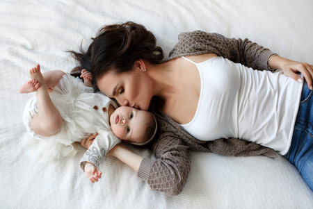 Mother and child on a white bed. Mom and baby boy in shorts playing in sunny bedroom. Parent and little kid relaxing at home.Family having fun together. Bedding and textile for infant nursery. Happy lfamily. Mother playing with her baby in the bedroomの写真素材