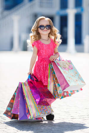 Beautiful happy smiling little girl child in sunglasses is holding shopping bags near shopping mall outdoors.Lifestyle concept.little shopaholic girl. girl with bags in hands.The delight of shopping.の写真素材