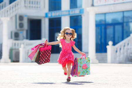 Beautiful happy smiling little girl child in sunglasses is holding shopping bags near shopping mall outdoors.Lifestyle concept.little shopaholic girl. girl with bags in hands.The delight of shopping.の写真素材