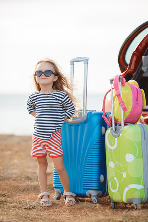 A little girl, a brunette with long curly hair, dressed in a striped sailor shirt, dark sun glasses, and a journey to the sea, stands near the open trunk of the red car with clothes, suitcases and bags.の写真素材