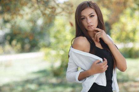 A beautiful young woman, a brunette with long hair and brown eyes, long black eyelashes and light make-up, dressed in a white shirt and black jersey, posing outdoors in the early autumn in the parkの写真素材
