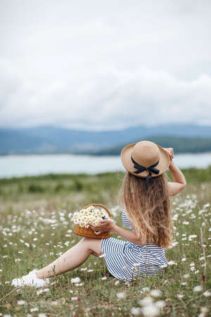 Happy woman having fun outdoor in nature.Young beautiful woman with blond long hair in a straw hat from the sun spends time alone outdoors in summer, sitting on a field among blooming white daisiesの写真素材