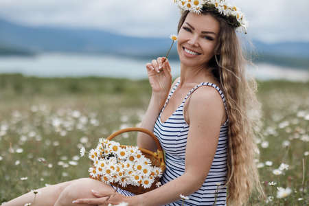 Young, beautiful woman in a wreath of wild flowers, spends time alone outdoors among white meadow daisies. Beautiful girl outdoors with a bouquet of flowers in a field of white daisies, enjoying nature.の写真素材