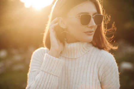 Natural beauty attractive young girl, portrait in a warm sweater in the park in early spring at sunset, sensual alluring look. A young beautiful woman in a white sweater walks alone in a spring parkの写真素材