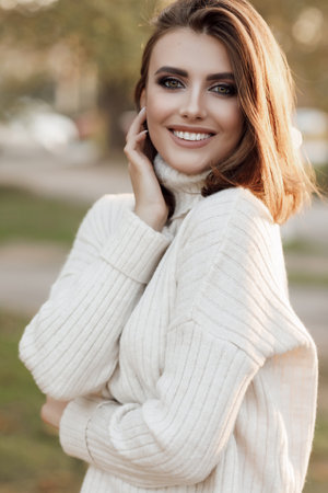 Natural beauty attractive young girl, portrait in a warm sweater in the park in early spring at sunset, sensual alluring look. A young beautiful woman in a white sweater walks alone in a spring parkの写真素材
