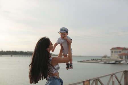 A beautiful young woman, a brunette with long straight hair, a sweet smile, plays with her young son outdoors in the summer on a wooden dais near the sea, holds the boy in his armsの写真素材