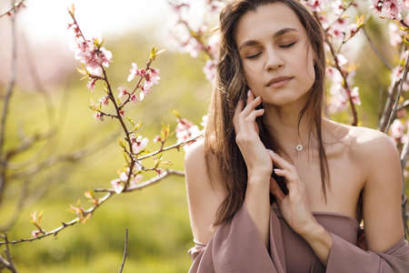 A beautiful young brunette with long hair in a light brown dress walks alone outdoors in a pink blooming peach garden in spring on a sunny day. Happy woman in the garden of blooming peaches.の写真素材
