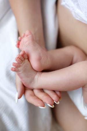 Newborn baby feet in mother hands. Close up of mother hand massaging foot newborn baby lying on comfortable white bed at home. Caucasian loving mom takes care of her infant son.の写真素材