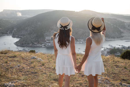 Two beautiful girls in white dresses and straw hats travel together in the highlands and enjoy the beautiful view of the quiet bay at sunset.の写真素材