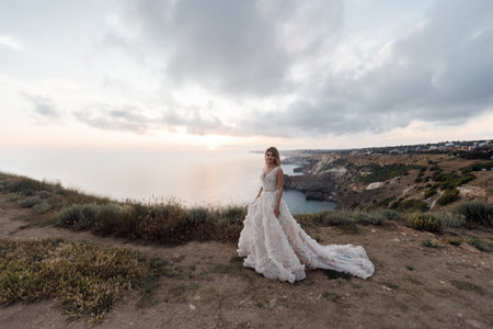 Bride in wedding dress poses while standing on a high steep bank against the backdrop of a quiet sea and clouds. Romantic bride in a luxurious dress posing on the beach in the evening at sunset.の写真素材