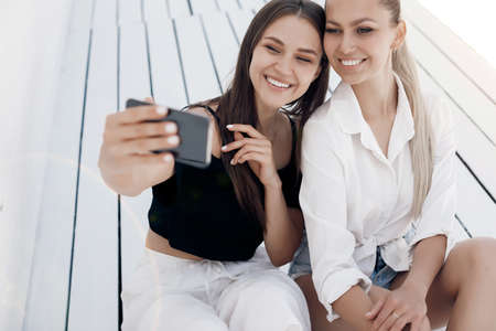 Beautiful girls spend time sitting on the beach of the mediterranean sea during summer vacation. Happy young women sitting on the seashore together on a white wooden deck rest and take a selfie.の写真素材