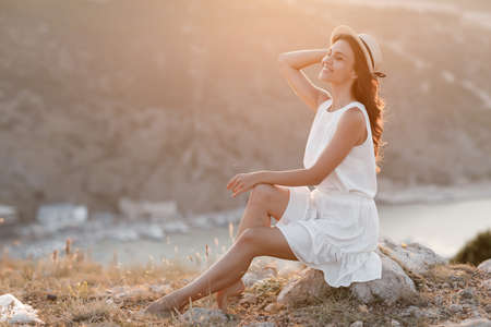 Happy young woman in white dress and straw hat is resting and relaxing sitting on a rocky hill and enjoying a beautiful view of a quiet bay in the evening at sunset.の写真素材