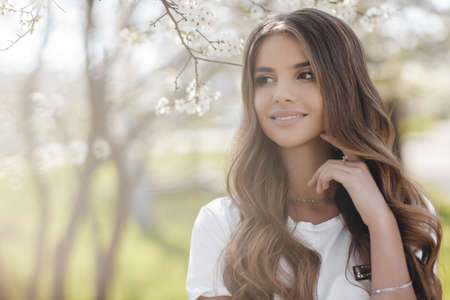 Young beautiful woman with a sweet smile in a blooming garden. A beautiful girl with long curly hair and brown eyes, in a white T-shirt, smiles and is happy, touching the flowers of a spring tree.の写真素材