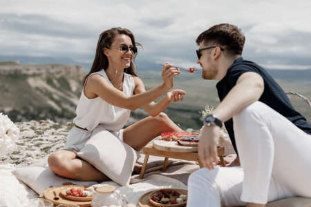 A loving couple at a romantic dinner in the mountains. Picnic for two on a high plateau. Young travelers sitting outdoors. Happy man and woman enjoying nature, relaxation, togetherness and travel.の写真素材