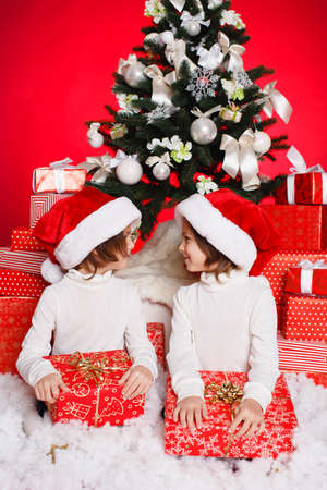 Little happy twinkling girls in red Santa Claus caps sit on the floor with gift boxes in front of a decorated Christmas tree, happy family celebrates Christmas and New Year.の写真素材