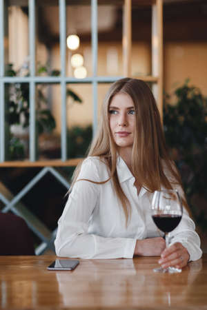 Young woman sitting alone at desk with glass of wine. Beautiful gorgeous girl drinking alcohol in a restaurant alone. Women's hands holding a large wine glass with red wine.の写真素材