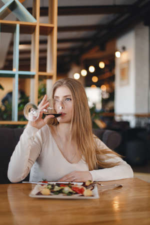 Young pretty woman with long hair dines with a glass of white wine in a restaurant. Inspired young woman enjoying champagne and salad in a restaurant on a summer weekend alone.の写真素材