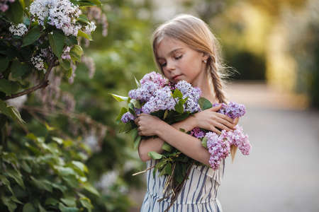 Spring portrait of a cheerful girl with a bouquet of lilac flowers in her hands. A beautiful girl, blonde, enjoys the aroma of blooming lilacs in the spring park, walking alone in the open air.の写真素材