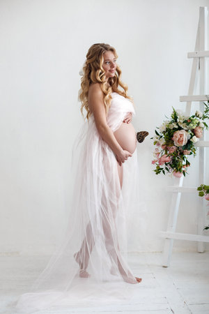 Partial nudity. Studio portrait of a pregnant woman with beautiful butterflies. Young pregnant woman with long blonde hair, while standing isolated on white studio background, expectant mother.の写真素材