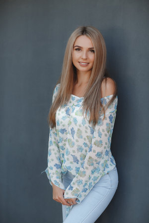 A beautiful young woman with long straight hair and brown eyes, a blonde with a pleasant smile, long black eyelashes dressed in a blue and white blouse, posing in the studio on a gray-blue backgroundの写真素材