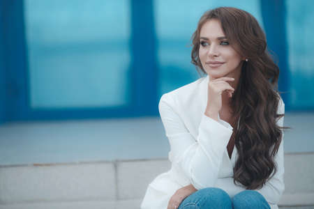 A young, beautiful, woman in the open air on a summer sunny day, gray big eyes and a sweet smile, long curly blond hair dressed in a stylish white jacket, spends time alone walking around the cityの写真素材