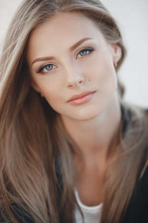 Closeup portrait of a young blonde girl with beautiful blue eyes wearing a black jacket in the open air. Pretty european woman with long straight hair. A woman in the city a white backgroundの写真素材