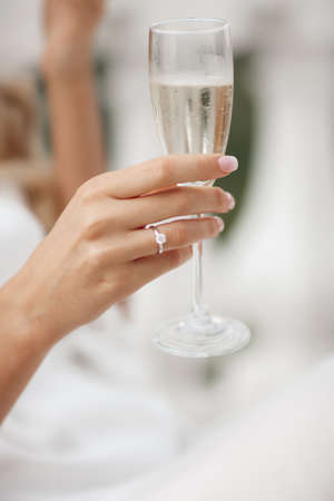 The bride listens to a wedding toast holding a glass of champagne in her hands. Hands of a young woman in a white wedding dress with a glass of white sparkling wine. Wishes of happinessの写真素材