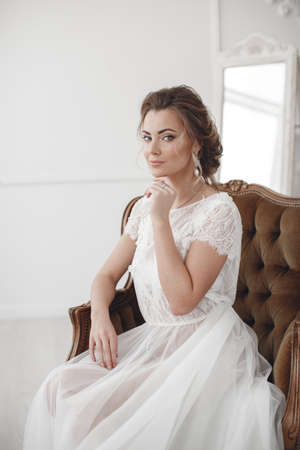Happy elegant young bride sitting in a chair.Pretty bride with wedding makeup,fashion jewelry.Beautiful woman posing in white wedding dress.Wedding portrait of happy bride.Gorgeous beauty,young brideの写真素材