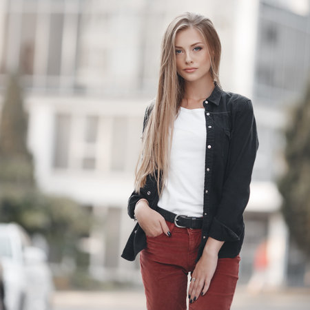 A young beautiful woman with long blond hair in a black jacket and brown trousers walks alone in the spring city on a sunny day.Stylish woman standing outside in front of big modern urban building.の写真素材