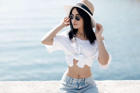 Summer portrait of a young beautiful brunette woman outdoors spending time alone posing by the sea in summer. A beautiful young woman in sunglasses resting on a promenade by the seの写真素材