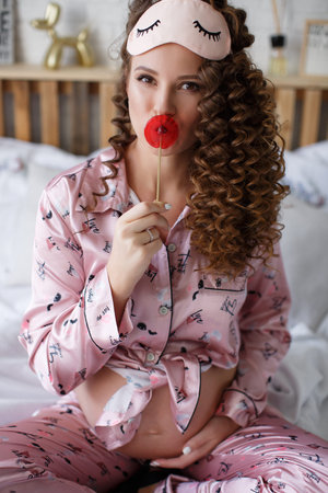 A beautiful young pregnant woman, a brunette with long thick curly hair, in pink pajamas, is sitting...