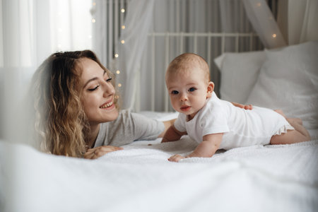 mother and child on a white bed. Mom and boy in a white t-shirt are playing in a sunny bedroom. Parent and small child rest at home. happy family. mother playing with her child.の写真素材