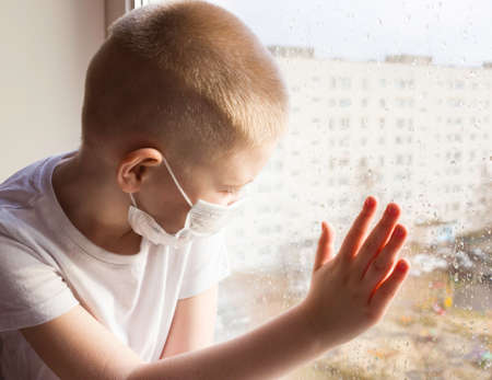 The concept of coronavirus, self-isolation, and quarantine. A boy in a disposable medical mask looks out the window with raindrops.の写真素材