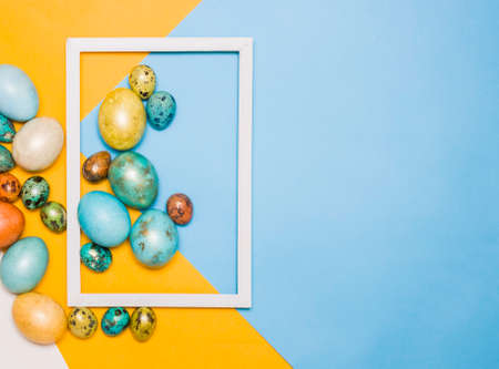 Painted Easter large and small eggs on a blue and yellow background, white wooden frame next to it. Free space for text. Happy Easter!の写真素材