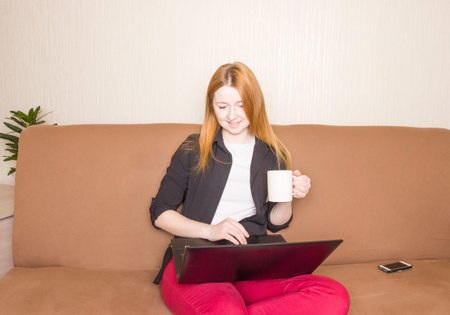 Distance learning online education and work. The girl is working on a laptop on the sofa and drinking coffee. Remote work.の写真素材