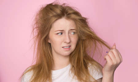 The concept of dry lifeless hair. A woman on a pink background holds her disheveled, tangled hair and looks at it with a discontented expression.の写真素材
