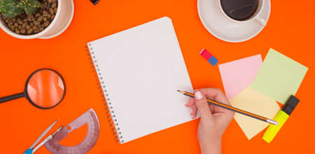 The concept of working in an office, mockup in a notebook. The hand holds a pencil and is about to write in an empty notebook. Stationery, coffee, and a flower. Top view, free space for text.の写真素材