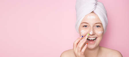 Smiling woman with a towel on her head on a pink background and face cream. Hydration of the skin, skin care face, beauty.の写真素材