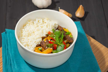 Roast beef with sweet pepper and white rice. White plate, garlic in the background, dark background.の写真素材
