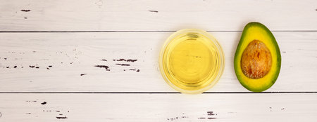 Avocado oil in a glass Cup and avocado cut with a stone on a white wooden background, top view, copyspace. Healthy food, vitamins, vegetarian food.の写真素材