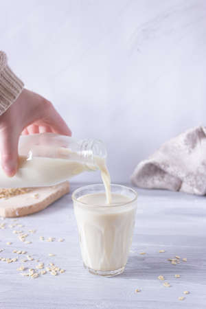 A hand pours vegetable oat milk from a bottle into a glass tumbler on a light background. Healthy drinks, vegetarianism. Side view. Vertical photoの写真素材